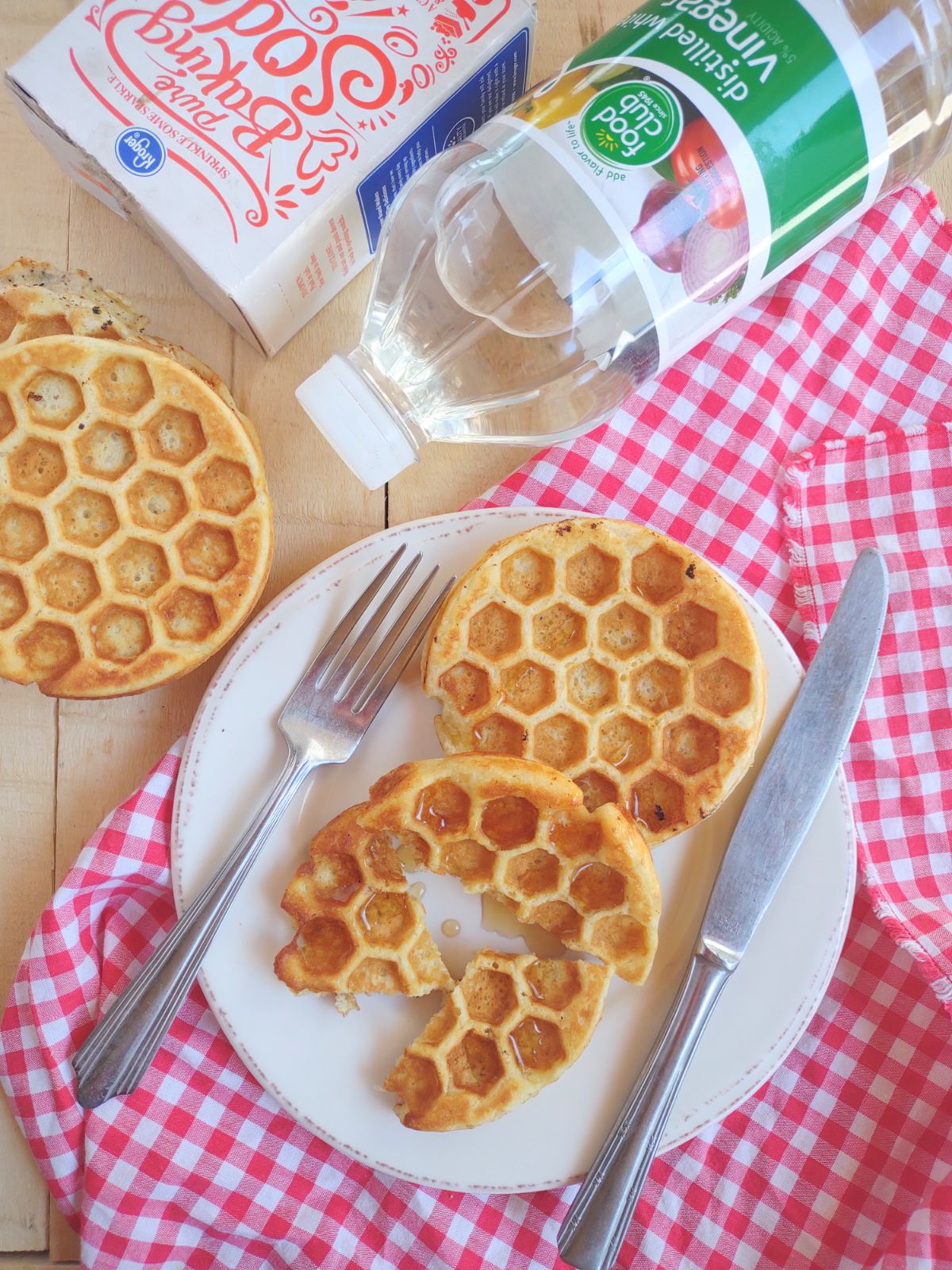Two waffles sitting on a white plate. One waffle is whole, the other one has been cut into pieces. The plate has a fork and knife on it and is sitting on a red plaid towel.