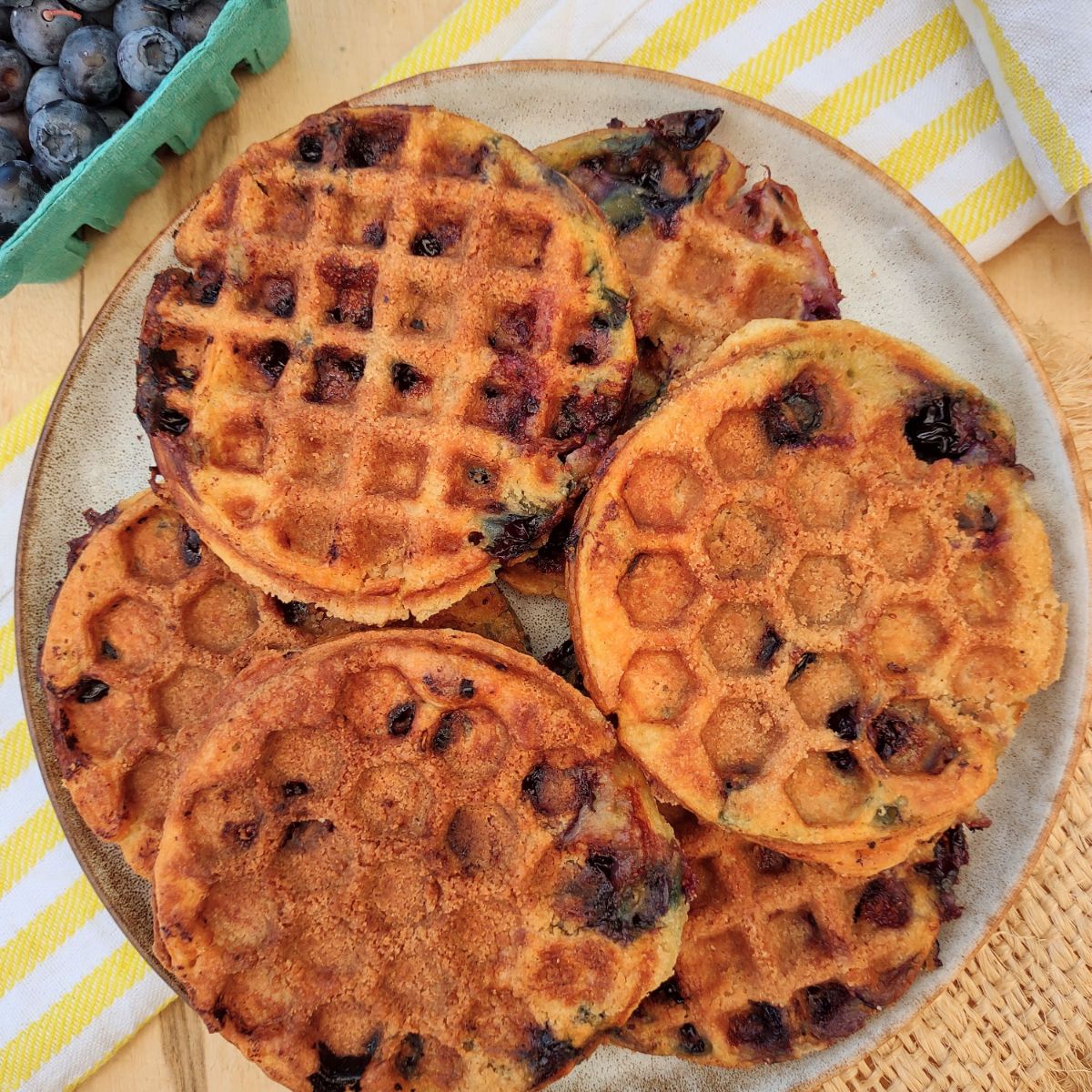Mini circle waffles that were cooked with a cobbler topping giving them a crunch exterior. The waffles are on a plate with a yellow striped towel underneath.