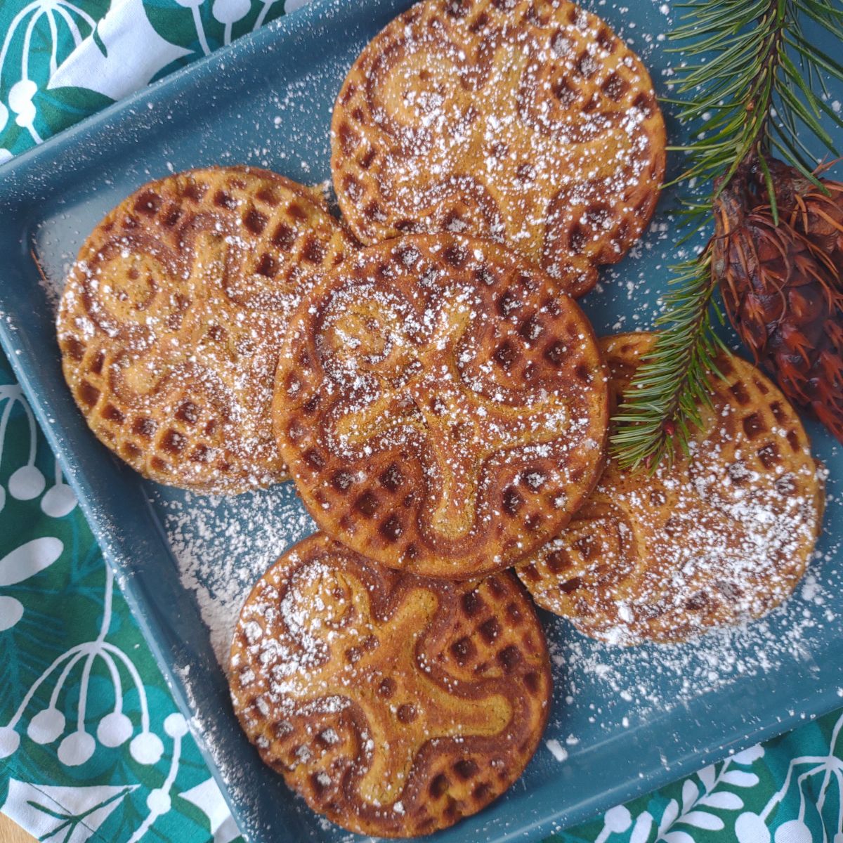 Gingerbread man waffles on a blue sheet pan topped with powdered sugar and a pine branch with cones attached next to them.
