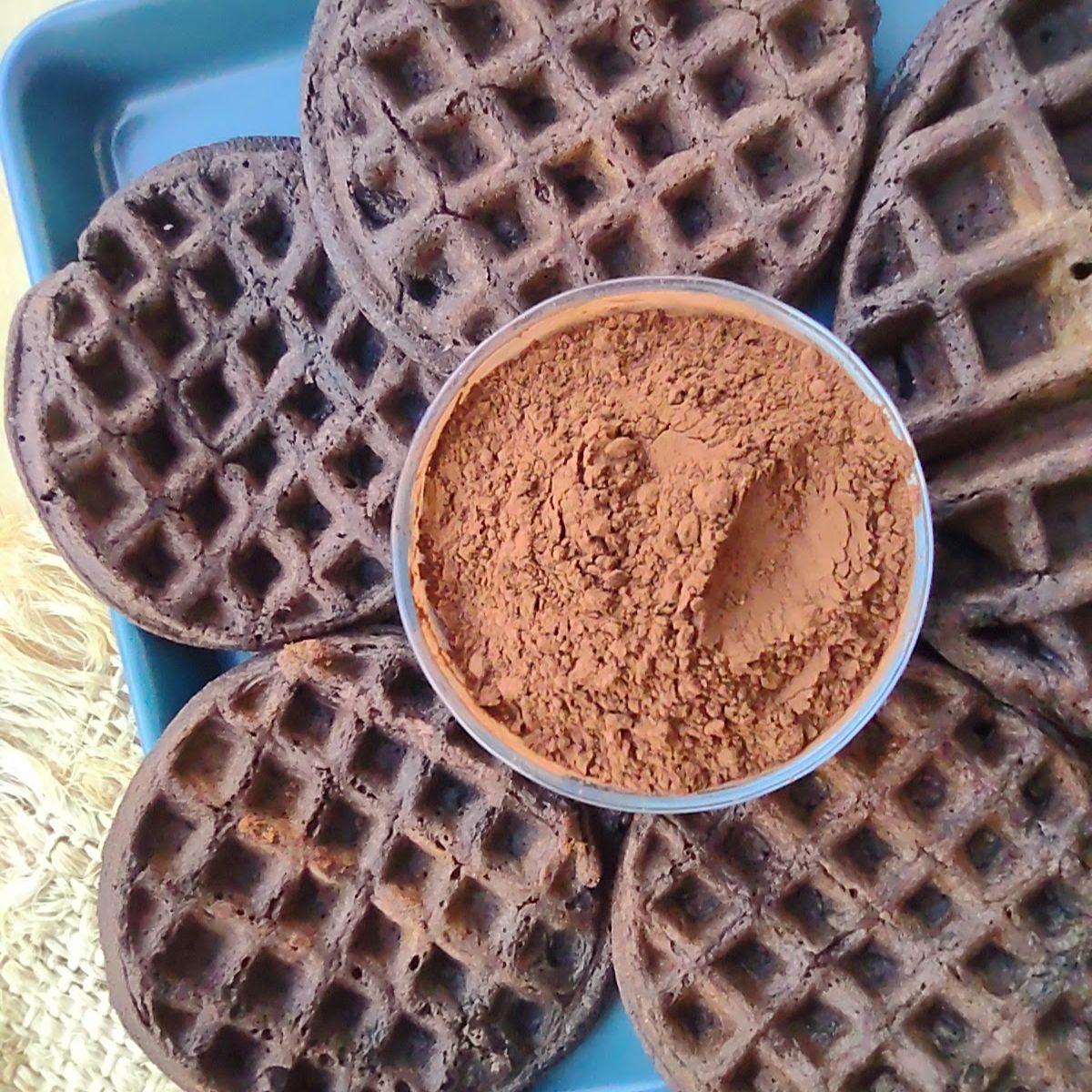 4 mini cocoa waffles and 1 large cocoa waffle surrounding a bowl of cocoa powder all on a blue sheet pan.