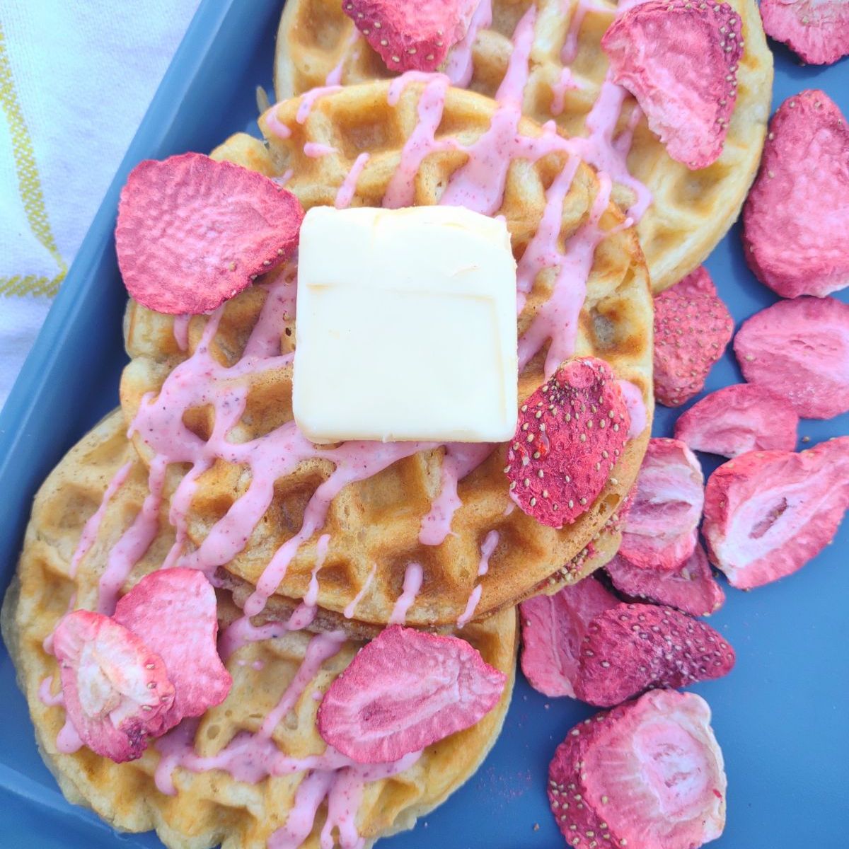 Circle mini waffles with a freeze dried strawberry icing drizzle and a pat of butter on a blue sheet pan.